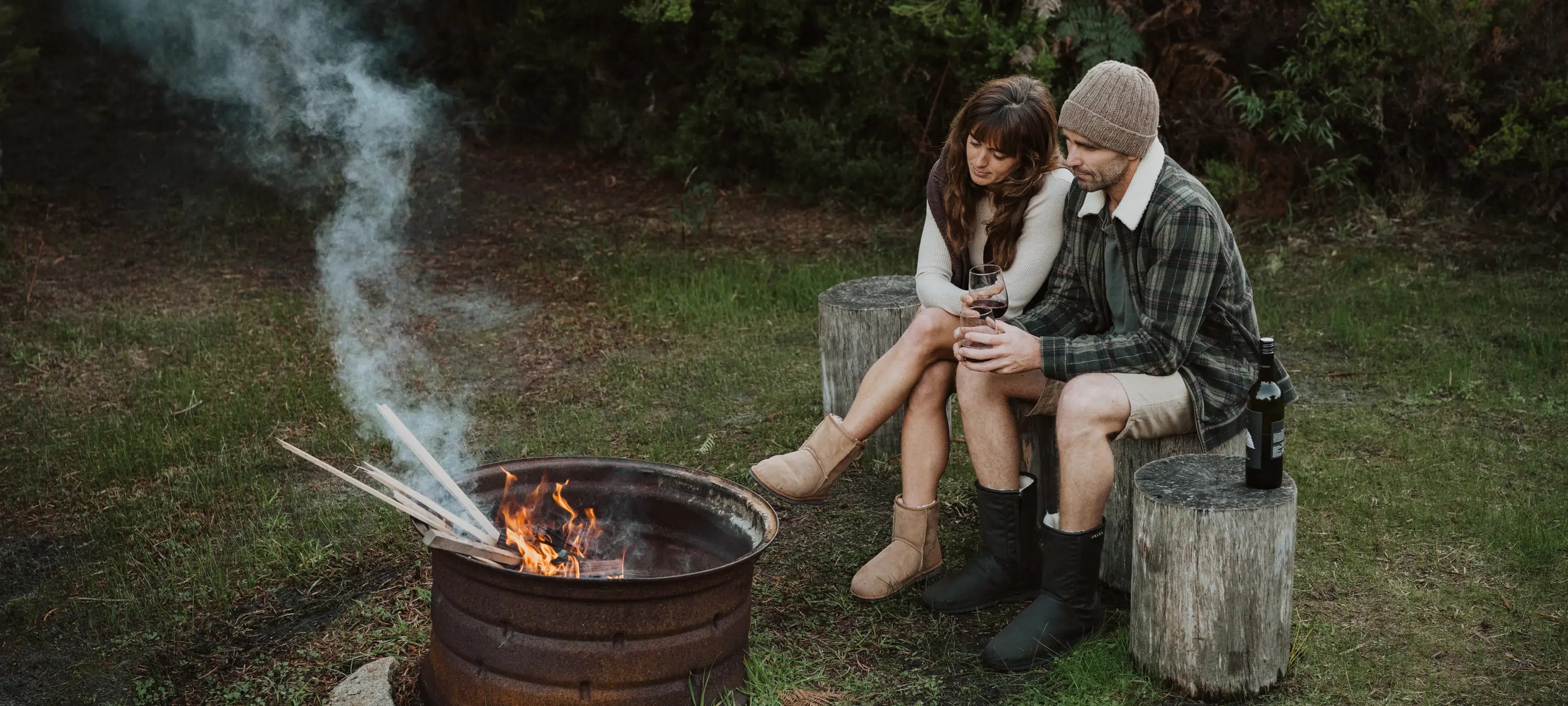 Two people wearing Jassi tan and oilskin Jassi ugg boots sitting by a campfire in a forest setting
