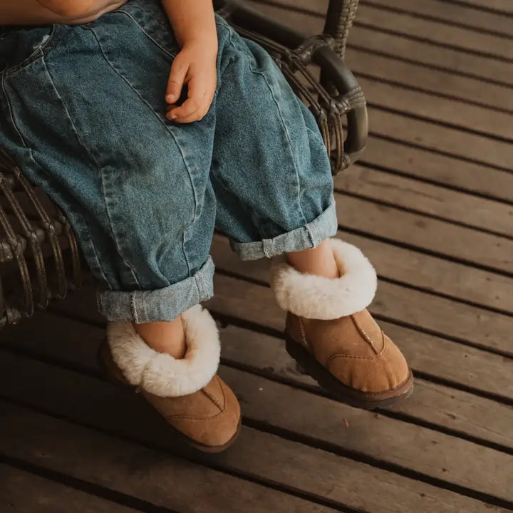 Close up of a kid sitting in a chair wearing Jassi toddler ugg boots in suede tan with a fluffy wool turn. #color_tan
