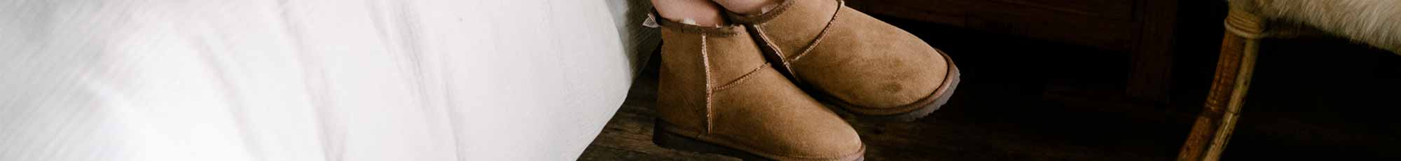 Short suede tan ugg boots hanging on the side of a bed.