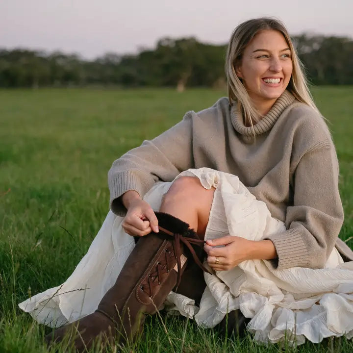 Woman sitting on a field wearing Jassi long lace-up UGG boots on suede chocolate. #color_chocolate