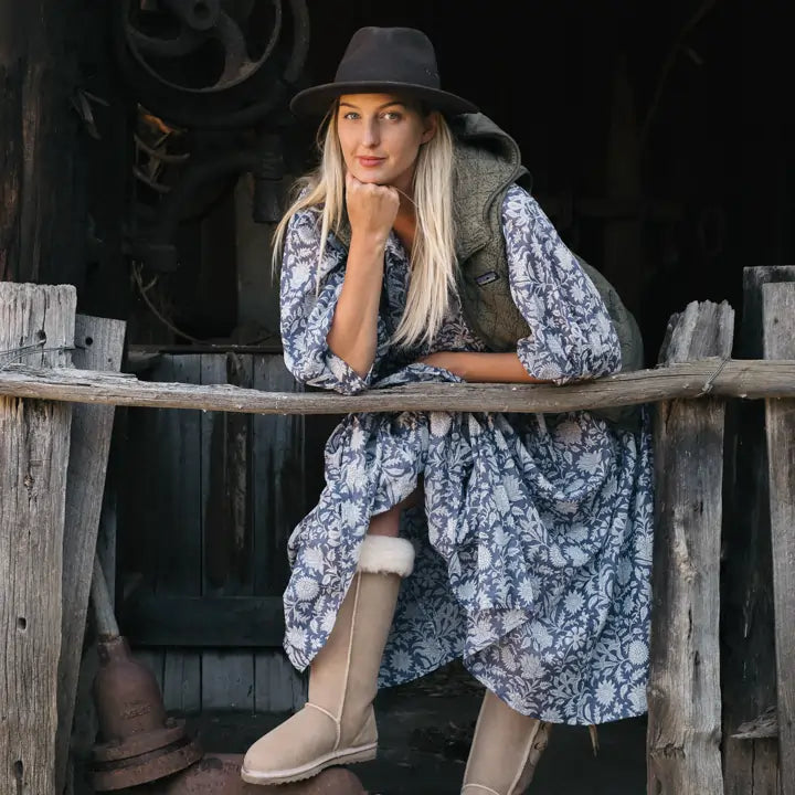 Woman in an old shed wearing a floral dress and Jassi Long Lace-up UGG Boots in sand suede. #color_sand