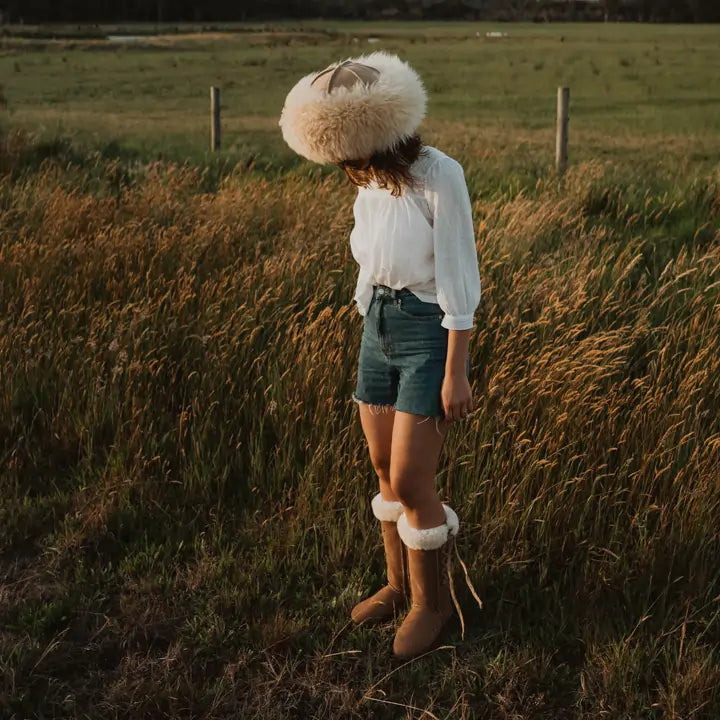 Woman on a field at sunset wearing a sheepskin hat and Jassi long lace-up UGG boots on suede tan. #color_tan