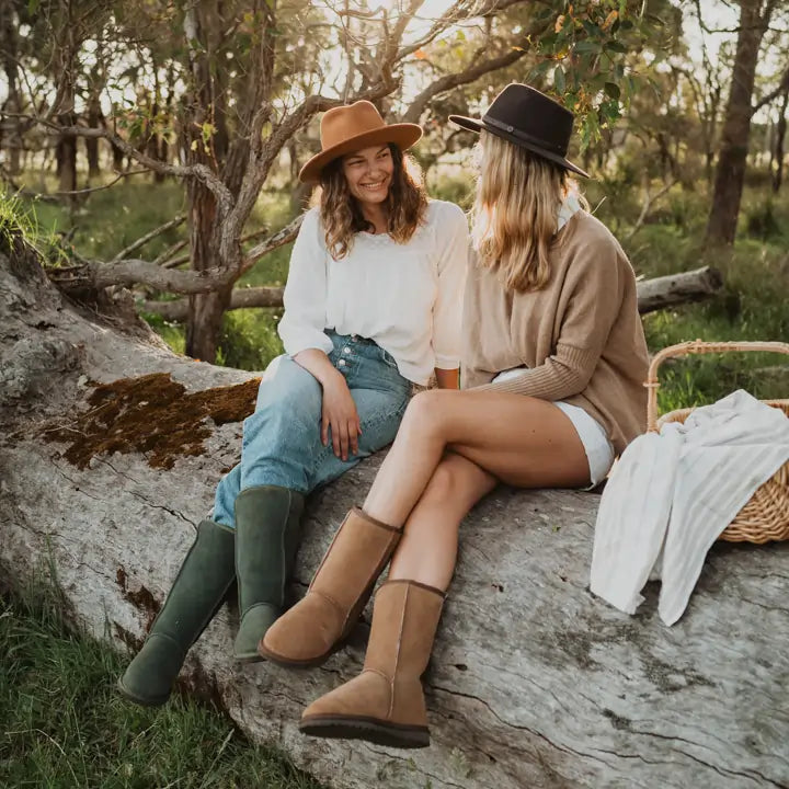 Two women sitting on a log with a picnic basket while wearing Jassi Long UGG Boots in olive and Jassi Mid UGG boots in suede tan. #color_olive