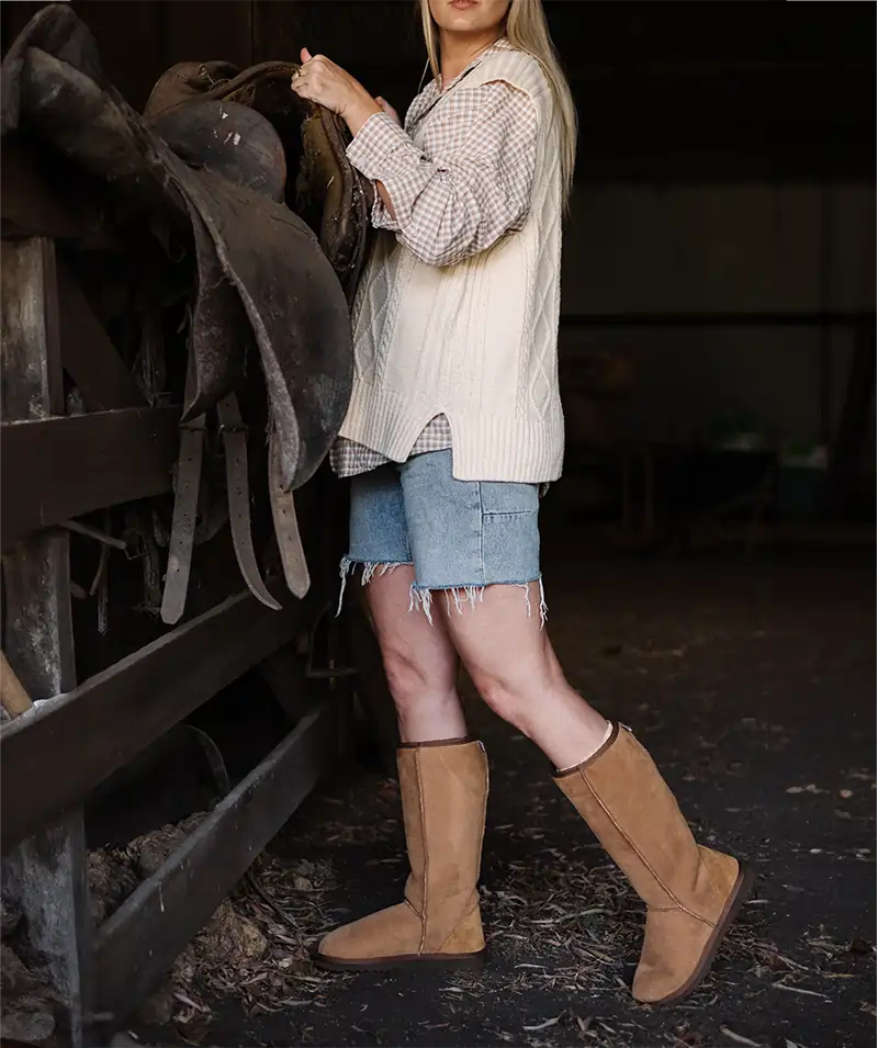 Woman picking up a saddle wearing Jassi long ugg boots in suede tan.