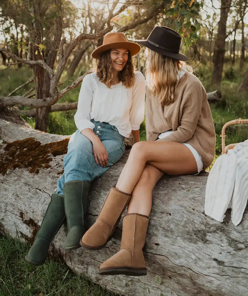 Two friends laughing while sitting on a log wearing Long ugg boots in olive color and Mid ugg boots in suede tan.