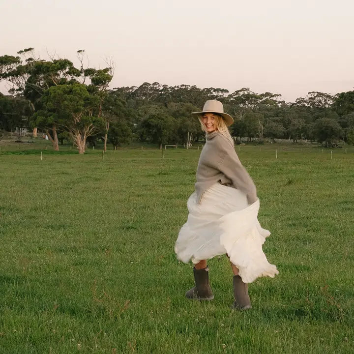 Woman running in a field wearing a white skirt and Jassi Mid UGG boots on grey suede. #color_grey