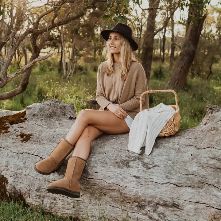 Woman sitting on a fallen log in the forest wearing Jassi mid UGG boots on suede tan color. #color_tan