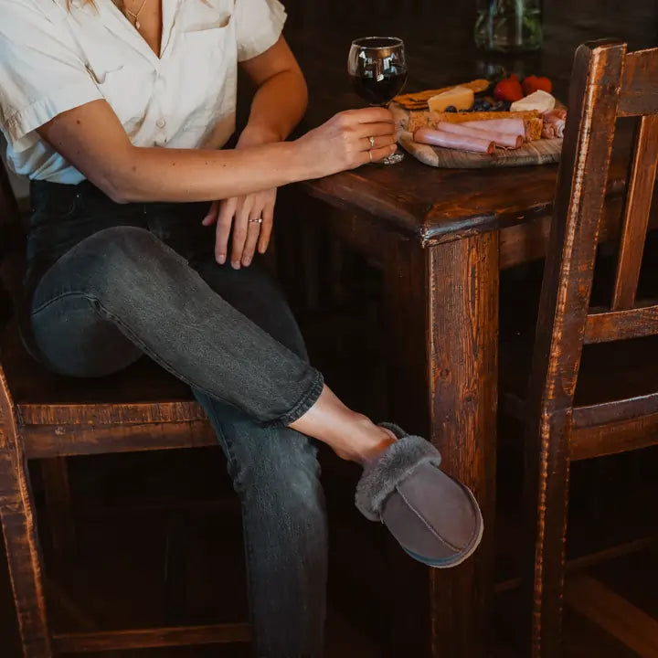 Woman having a wine while wearing Jassi ugg scuffs in grey suede with a wool turn. #color_grey