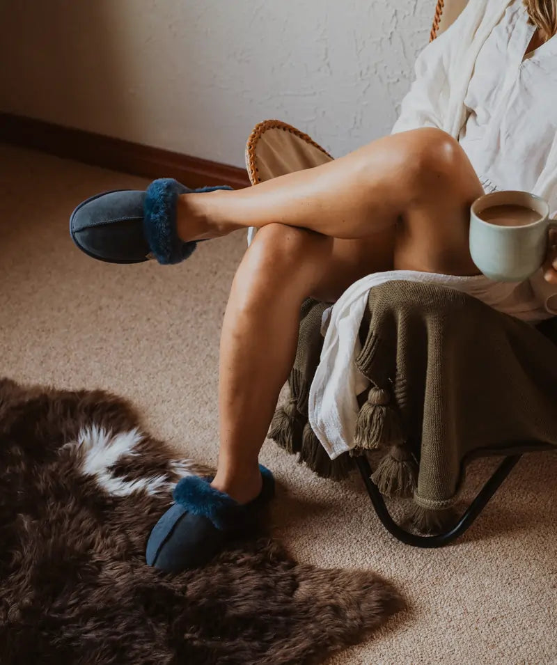 Woman having a cup of tea while wearing Jassi ugg scuffs in suede finish and navy color.