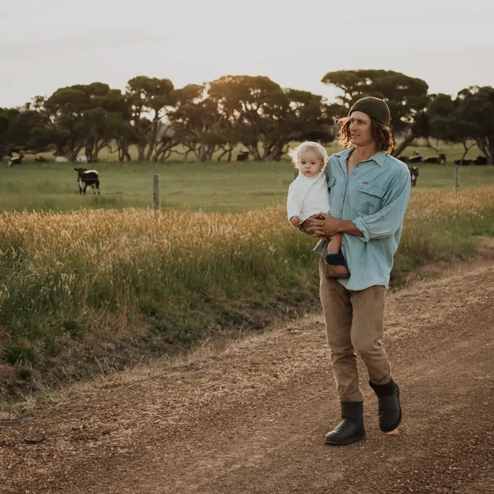 Man carrying a baby on a farm while wearing Jassi leather short ugg boots in chocolate color. #color_leather chocolate