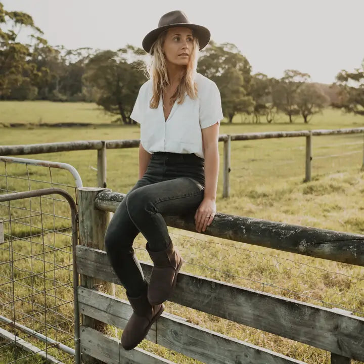 Woman sitting on a farm fence wearing Jassi short UGG boots in brown suede color. #color_chocolate