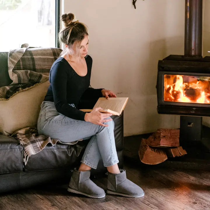 Woman reading a book next to a fireplace while wearing Jassi Short UGG Boots in suede grey. #color_grey