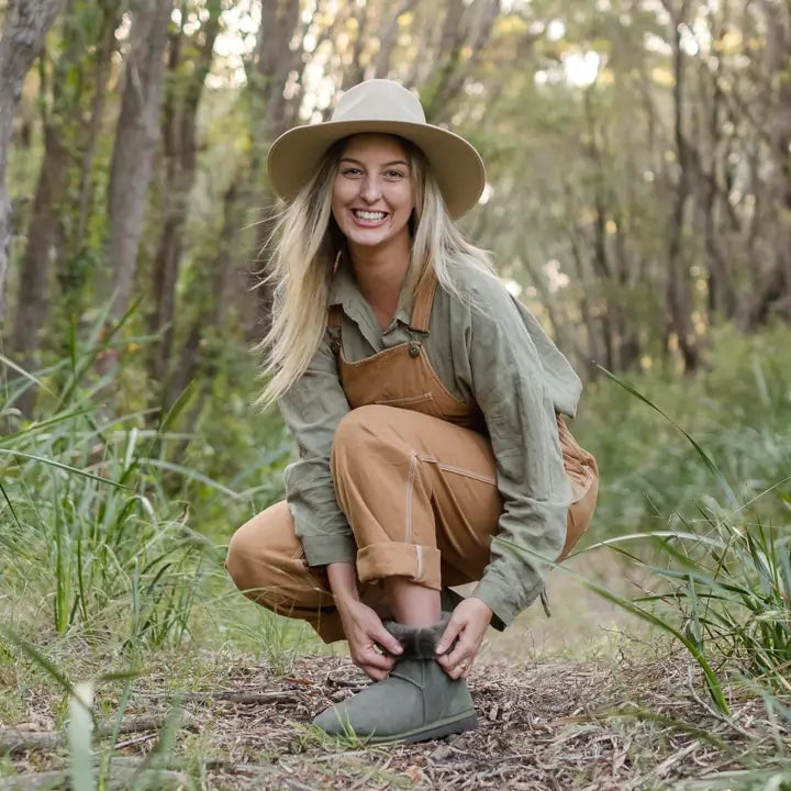 Smiling young woman adjusting her short suede olive ugg boots in a forest. #color_olive