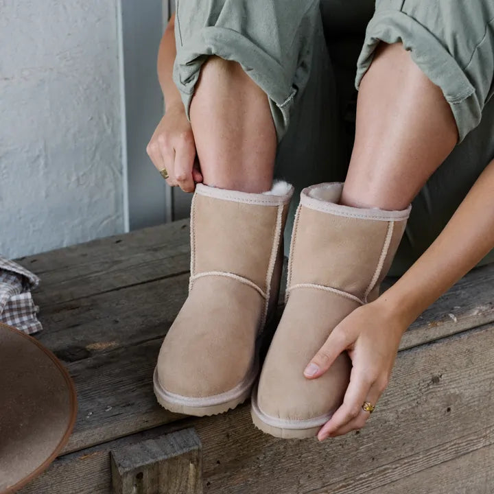 Woman sitting in a bench wearing cosy Jassi Short UGG Boots in suede sand color. #color_sand