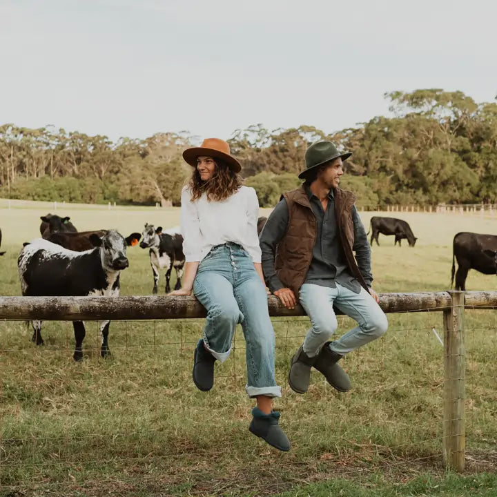 Man and a woman sitting on a farm fence with cows on the background wearing Jassi ugg slippers in navy color. #color_navy