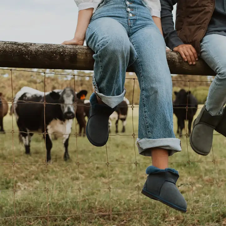 Close up of woman sitting on a farm fence wearing Jassi ugg slippers in navy color. #color_navy