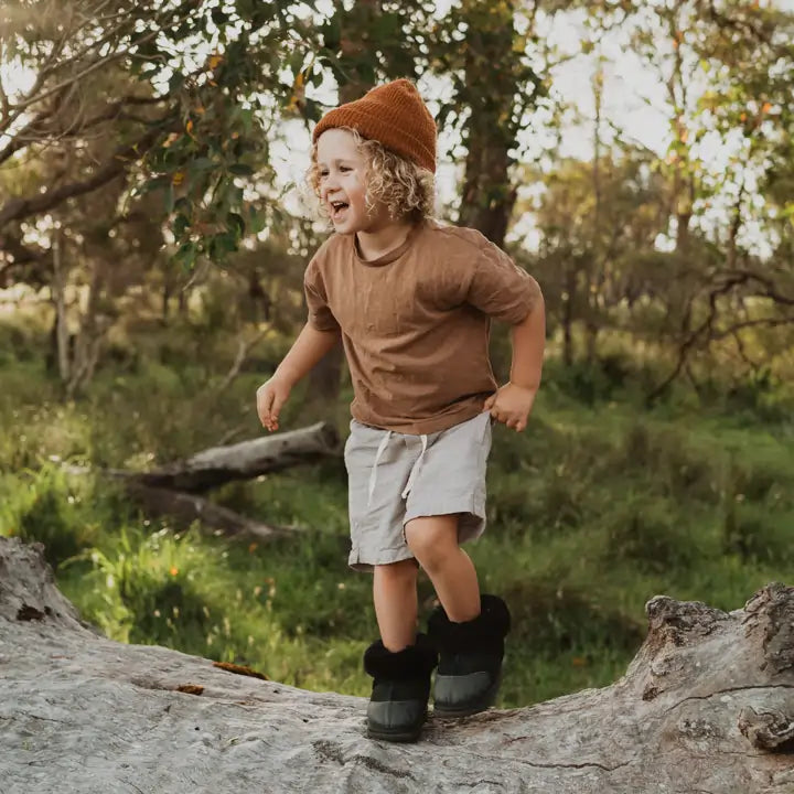 Little kid playing on top of a fallen tree wearing Jassi toddler ugg boots in suede black with a leather reinforcement. #color_black