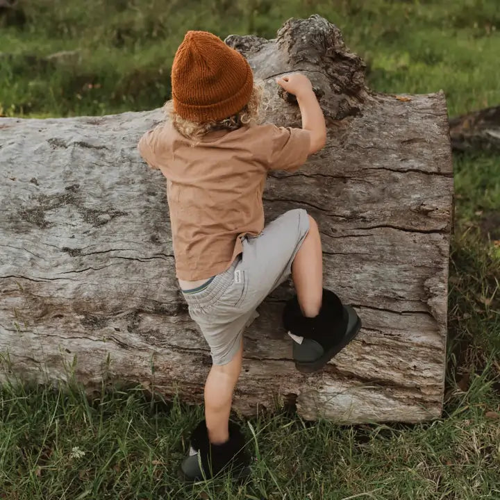 Little kid climbing a fallen tree wearing Jassi toddler ugg boots in suede black.  #color_black