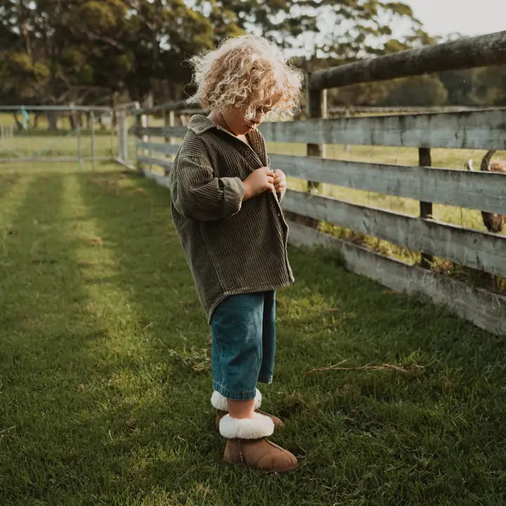 Little kid buttoning his shirt in a farm wearing Jassi toddler ugg boots in suede tan. #color_tan